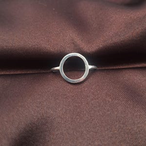 Puede incluir: Un anillo de plata con un diseño minimalista con un centro abierto circular. El anillo está colocado sobre un fondo de tela marrón, resaltando su elegante sencillez. Esta joya es adecuada para el uso diario.