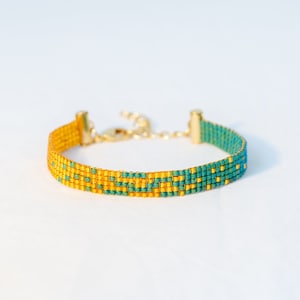 May include: A beaded bracelet with a gold clasp and chain. The bracelet features a pattern of teal and gold beads. The bracelet is on a white background.