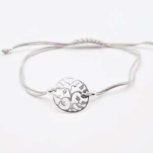 Puede incluir: Un brazalete plateado con un dije de árbol de la vida en un cordón gris.