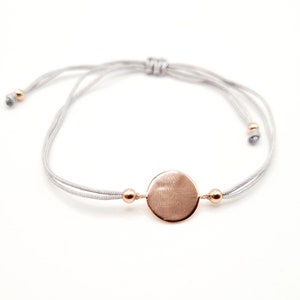 Könnte beinhalten: Ein verstellbares Armband aus grauem Kordel mit einem runden Anhänger aus Roségold und kleinen Perlen aus Roségold.