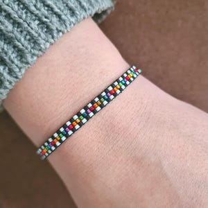 Può includere: Un braccialetto di perline con base nera e un motivo di perline bianche, arcobaleno e nere. Il braccialetto è indossato su un polso, con una manica di maglione grigia visibile sullo sfondo. Il braccialetto è largo circa 1,3 cm.