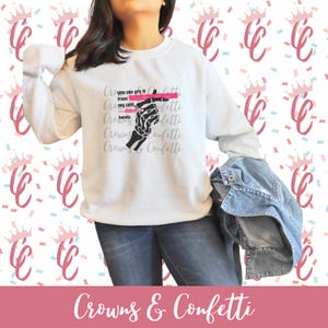 Può includere: Felpa bianca con grafica di una mano scheletrica e la scritta "you can pry it from my cold, dead hands". Le parole "Crowns & Confetti" sono in basso nell'immagine. Lo sfondo presenta un motivo ripetuto di corone e coriandoli.