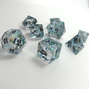 Puede incluir: Un conjunto de siete dados iridiscentes azules y verdes con números negros. Los dados están hechos de resina transparente con purpurina incrustada en la resina. Los dados están dispersos sobre una superficie blanca.