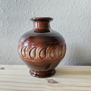 May include: A small, round ceramic vase with a glossy, deep brown glaze. The body of the vase features a horizontal band of embossed, teardrop-shaped patterns encircling the center. It has a short, narrow neck and a flared base rim.