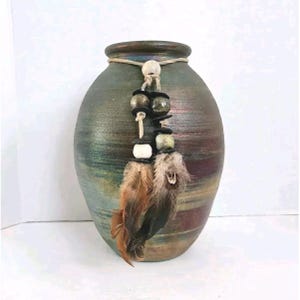 May include: A rounded ceramic vase with a textured, multi-colored finish in shades of green, brown, and blue. A decorative cord is tied around the neck, featuring wooden beads and two dangling brown and black feathers hanging down the front.