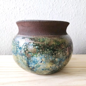 May include: A small, round ceramic pot featuring a dark brown, matte-textured rim and a bulbous body. The lower section displays a mottled glaze with blended shades of deep green, earthy brown, and vibrant blue, creating a textured appearance.