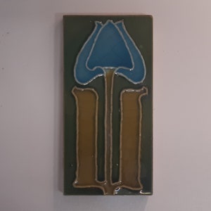 Puede incluir: Azulejo cerámico rectangular con un diseño floral estilizado. La flor azul tiene un contorno plateado, sobre un fondo verde. Dos formas verticales marrones forman el tallo. La baldosa es probablemente para uso decorativo.