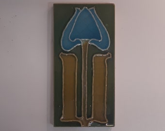 May include: Rectangular ceramic tile with a stylized floral design. The flower is blue with a silver outline, set against a green background. Two brown vertical shapes form the stem. The tile is likely for decorative use.