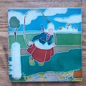 May include: A vintage ceramic tile depicting a girl in a red and blue dress jumping rope in a green field. The tile has a blue sky with white clouds and a small white church in the background.