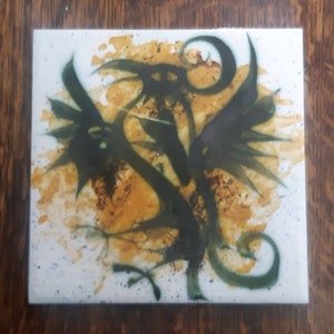 May include: A white ceramic tile with a green and yellow abstract design. The design features two stylised birds with wings spread wide. The background is a light yellow with white speckles.
