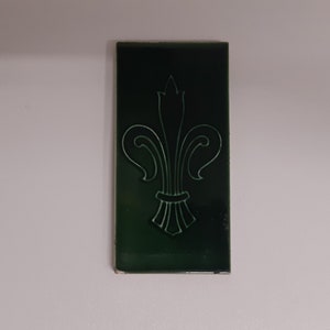 May include: A dark green ceramic tile with a white fleur-de-lis design.