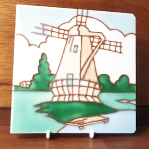May include: Square ceramic tile depicting a windmill scene. The windmill is beige with brown detailing, set against a light blue sky with white clouds. Green trees and water complete the landscape. The tile is displayed on a white stand.