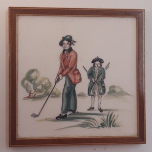 Könnte beinhalten: Eine handbemalte Fliese, die zwei Figuren in Kleidung aus dem 18. Jahrhundert beim Golfspielen zeigt. Die Figur im Vordergrund schwingt einen Golfschläger, während die andere Figur mit einem Schläger in der Hand dahinter steht. Die Fliese hat einen weißen Hintergrund mit grünen und braunen Akzenten.