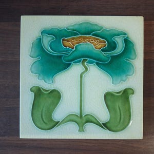 May include: A square ceramic tile featuring a raised relief design of a stylised flower. The flower has vibrant teal petals, a golden-brown centre, and two large green leaves at the base. The background is a pale cream colour with a fine crackle glaze.