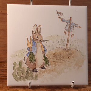 Könnte beinhalten: Eine handbemalte Keramikfliese mit einer Aquarell-Illustration von Peter Rabbit, der Karotten hält, und einem Vogelscheuchen im Hintergrund.
