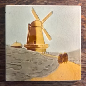 May include: Square ceramic tile depicting a windmill scene. The windmill is a warm yellow-orange colour with four blades. The background is a light grey, and the foreground is a sandy yellow-orange. The tile has a slightly aged appearance.