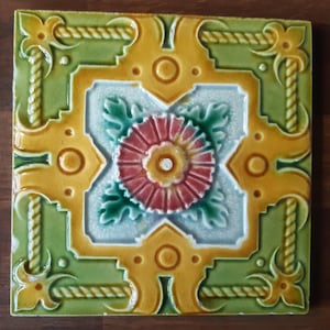 May include: Square ceramic tile with a floral design. The tile features a central red flower with a yellow center, surrounded by green leaves. The border is green and gold, with rope-like detailing and decorative corner elements.