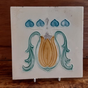 May include: A white ceramic tile with a decorative design featuring a stylised yellow flower with blue and green accents.