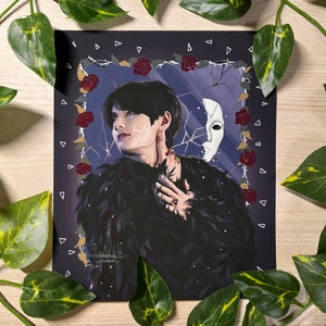 May include: A digital illustration of a person with dark hair wearing a black feathered jacket and a ring on their finger. The person is looking to the side and has a white mask on their face. The background is dark blue with white cracks and red roses.