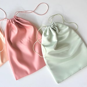 May include: Three satin drawstring bags in pastel colors: peach, rose, and mint green. Each bag has a matching drawstring closure. The bags are arranged on a white surface, showcasing their smooth, shiny texture.
