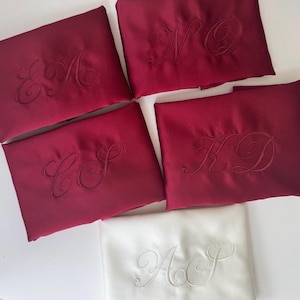 Pode incluir: Quatro bolsas retangulares de cetim bord&ocirc; e uma bolsa de cetim branco, cada uma com um monograma bordado tom sobre tom em letra cursiva. As bolsas bord&ocirc; exibem as letras EA, NQ, CS e KD, enquanto a bolsa branca exibe as letras AS.