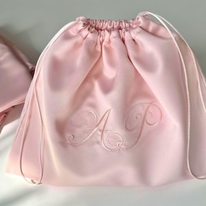 Peut inclure: Une pochette à cordon en satin rose clair avec une finition lisse et brillante, dotée d'une fermeture froncée avec des cordons assortis. Le centre de la pochette est brodé des lettres monogrammes A et P en cursive avec un fil ton sur ton.