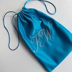 Pode incluir: Uma bolsa com cord&atilde;o de cor turquesa brilhante feita de um tecido liso e acetinado. A parte frontal da bolsa apresenta as letras KW bordadas numa fonte cursiva branca. A bolsa &eacute; fechada no topo com cord&otilde;es turquesa a condizer.