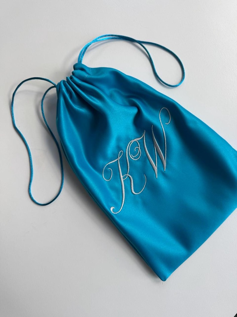 Pode incluir: Uma bolsa com cord&atilde;o de cor turquesa brilhante feita de um tecido liso e acetinado. A parte frontal da bolsa apresenta as letras KW bordadas numa fonte cursiva branca. A bolsa &eacute; fechada no topo com cord&otilde;es turquesa a condizer.