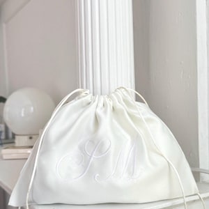 Pode incluir: Uma bolsa de cetim branco com cord&atilde;o, apresentando um delicado monograma bordado tom sobre tom com as letras SM em letra cursiva. A bolsa &eacute; franzida no topo com cord&otilde;es de cetim branco a condizer, criando uma silhueta suave e franzida.