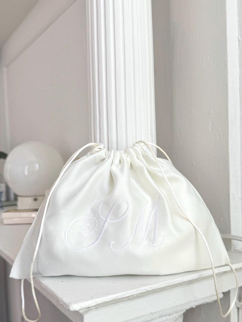 Pode incluir: Uma bolsa de cetim branco com cord&atilde;o, apresentando um delicado monograma bordado tom sobre tom com as letras SM em letra cursiva. A bolsa &eacute; franzida no topo com cord&otilde;es de cetim branco a condizer, criando uma silhueta suave e franzida.