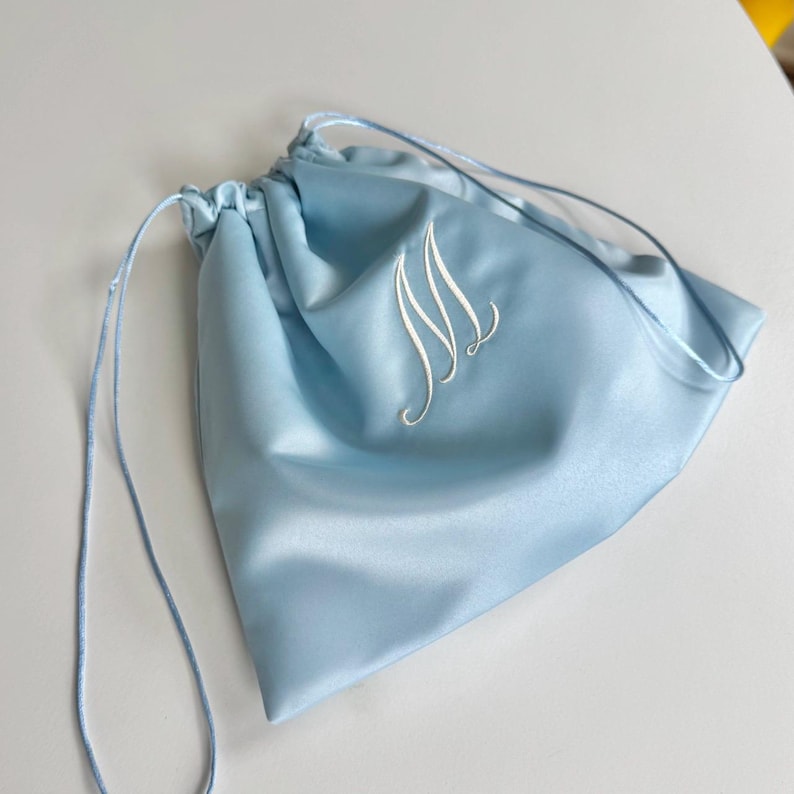 Pode incluir: Uma bolsa de cetim azul claro com cord&atilde;o, apresentando uma letra M bordada em branco em fonte cursiva. A bolsa possui cord&otilde;es de tecido azul claro correspondentes em ambos os lados, criando um fechamento franzido para este acess&oacute;rio de armazenamento.