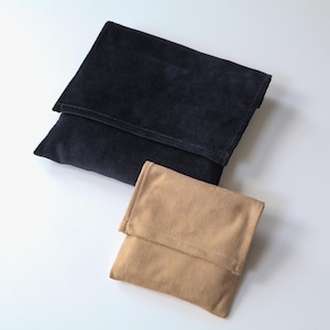 Puede incluir: Dos bolsos de ante, uno azul marino y otro beige, se muestran sobre una superficie blanca. Los bolsos tienen forma rectangular con un cierre de solapa plegada. El bolso azul marino es más grande que el beige.