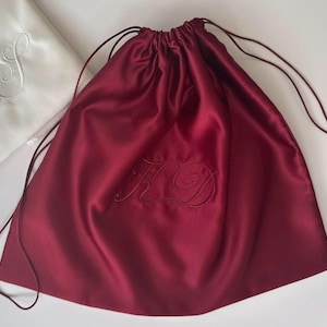 Pode incluir: Uma bolsa de cetim bord&ocirc; profundo com cord&atilde;o e as iniciais KD bordadas em letra cursiva. Ao lado, h&aacute; uma bolsa de cetim branca com as iniciais AS bordadas em cursivo correspondente, ambas com cord&otilde;es finos e combinando.