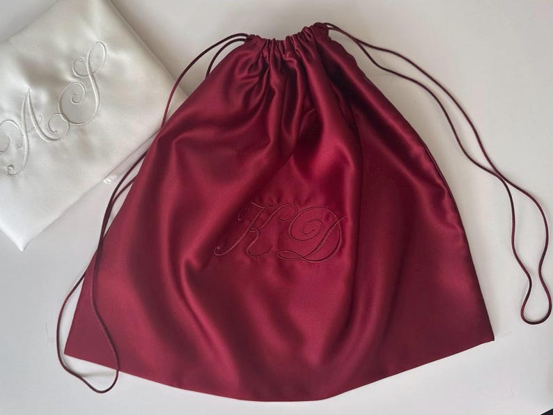 Pode incluir: Uma bolsa de cetim bord&ocirc; profundo com cord&atilde;o e as iniciais KD bordadas em letra cursiva. Ao lado, h&aacute; uma bolsa de cetim branca com as iniciais AS bordadas em cursivo correspondente, ambas com cord&otilde;es finos e combinando.