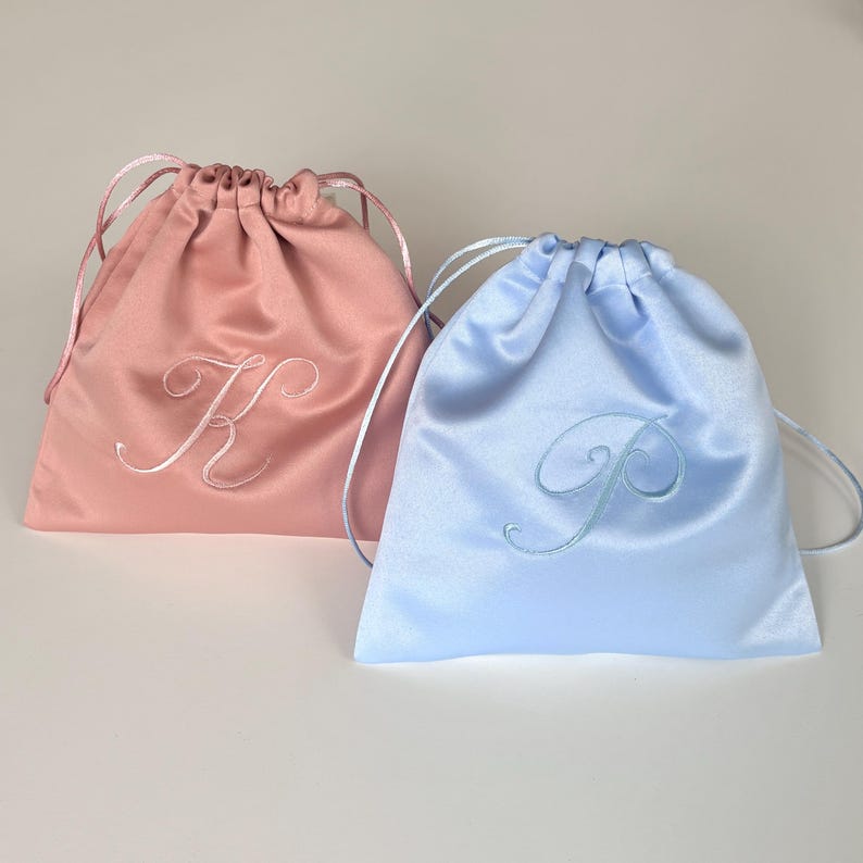 Pode incluir: Dois sacos de cetim com cord&atilde;o, um em rosa seco e outro em azul claro. Cada saco apresenta uma letra bordada em escrita cursiva na parte frontal, a letra K no saco rosa e a letra P no azul, com fitas a condizer para o fecho.