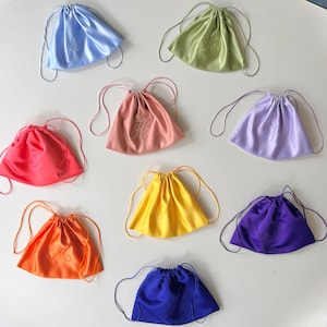 Pode incluir: Uma cole&ccedil;&atilde;o de nove bolsas com cord&atilde;o feitas de tecido liso semelhante a cetim em v&aacute;rias cores, incluindo azul claro, verde s&aacute;lvia, rosa, lavanda, vermelho brilhante, amarelo, laranja, roxo e azul royal, cada uma com uma letra bordada.