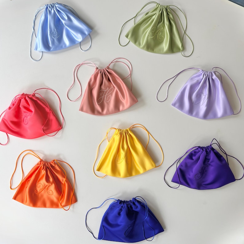 Pode incluir: Uma cole&ccedil;&atilde;o de nove bolsas com cord&atilde;o feitas de tecido liso semelhante a cetim em v&aacute;rias cores, incluindo azul claro, verde s&aacute;lvia, rosa, lavanda, vermelho brilhante, amarelo, laranja, roxo e azul royal, cada uma com uma letra bordada.