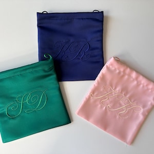 Pode incluir: Tr&ecirc;s bolsas quadradas de cetim com cord&atilde;o nas cores azul-marinho, verde-esmeralda e rosa-claro. Cada bolsa apresenta um monograma bordado tom sobre tom em letra cursiva ao centro. A bolsa azul mostra KR, a verde SD e a rosa KH.
