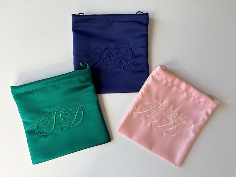 Pode incluir: Tr&ecirc;s bolsas quadradas de cetim com cord&atilde;o nas cores azul-marinho, verde-esmeralda e rosa-claro. Cada bolsa apresenta um monograma bordado tom sobre tom em letra cursiva ao centro. A bolsa azul mostra KR, a verde SD e a rosa KH.