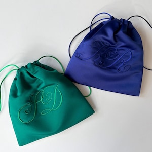 Pode incluir: Dois sacos com cord&atilde;o feitos de tecido liso, um em verde esmeralda e outro em azul real. Cada saco apresenta um monograma bordado a condizer com letras cursivas na frente e cord&otilde;es finos para fechar e transportar.
