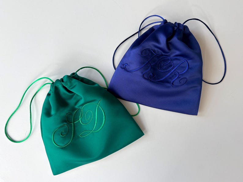 Pode incluir: Dois sacos com cord&atilde;o feitos de tecido liso, um em verde esmeralda e outro em azul real. Cada saco apresenta um monograma bordado a condizer com letras cursivas na frente e cord&otilde;es finos para fechar e transportar.