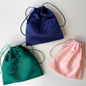 Pode incluir: Tr&ecirc;s bolsas de cetim com cord&atilde;o nas cores azul-marinho, verde-esmeralda e rosa claro. Cada bolsa apresenta um monograma bordado no mesmo tom na parte frontal e inclui cord&otilde;es de cetim da mesma cor para fechar no topo.