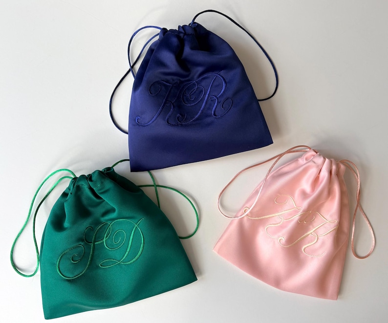 Pode incluir: Tr&ecirc;s bolsas de cetim com cord&atilde;o nas cores azul-marinho, verde-esmeralda e rosa claro. Cada bolsa apresenta um monograma bordado no mesmo tom na parte frontal e inclui cord&otilde;es de cetim da mesma cor para fechar no topo.