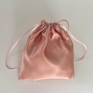 Pode incluir: Uma bolsa de cordão de cetim rosa claro com um cordão branco.