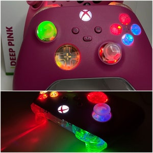 May include: A pink Xbox controller with colorful lights on the buttons and the Xbox logo. The controller is on a white surface with the words "DEEP PINK" visible.