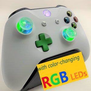 May include: White Xbox controller with green buttons and two clear, color-changing RGB LED thumbsticks. The text "with color-changing RGB LEDs" is on a yellow background.