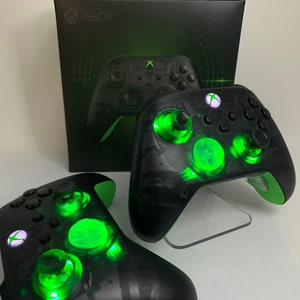 以下が含まれることがあります： 緑色の光るボタンと半透明の黒いデザインが施された黒いXboxコントローラーが2つ。コントローラーは白い表面に置かれており、背景には「XBOX」と書かれた黒い箱がある。