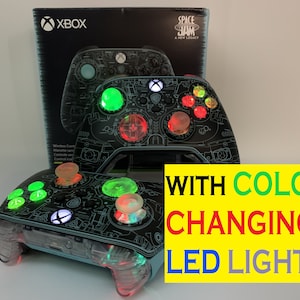 May include: A black and blue Xbox controller with a Space Jam: A New Legacy design. The controller has color-changing LED lights on the buttons and the Xbox logo.
