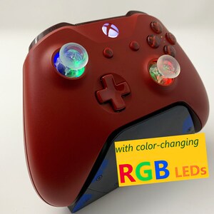 May include: Red Xbox controller with color-changing RGB LEDs on the thumbsticks. The controller is on a charging stand.