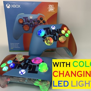 May include: Two blue Xbox controllers with a "Tune Squad" design and color-changing LED lights. The controllers are on a white surface with a box in the background.